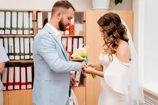 Bride in a stylish white dress holding a small bouquet outside a city hall building
