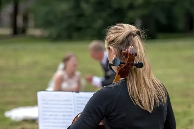 Cellist performing outdoors while a couple in formal attire sits on the grass in the background